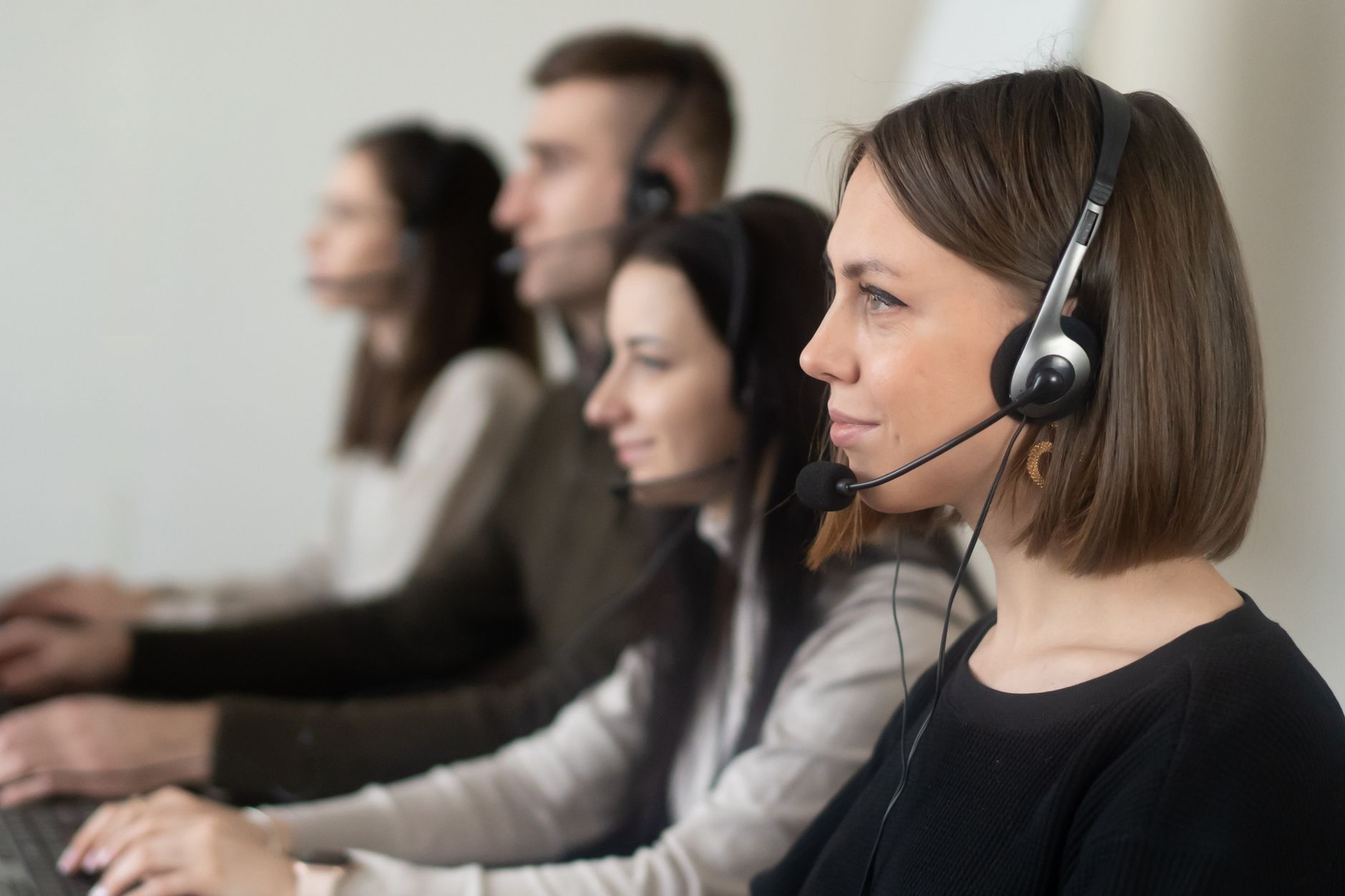 Side view of young coworkers in casual clothes sitting in light workplace with headphones with microphone while surfing on laptop